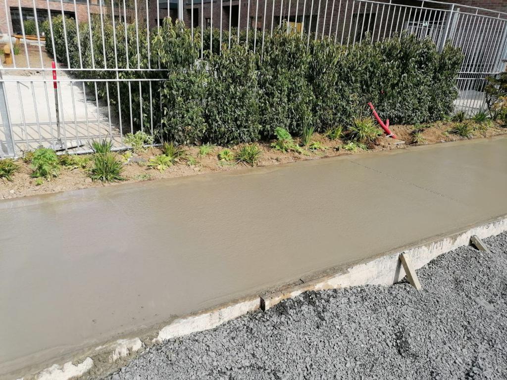 wet concrete sidewalk under construction with a metal fence and bushes